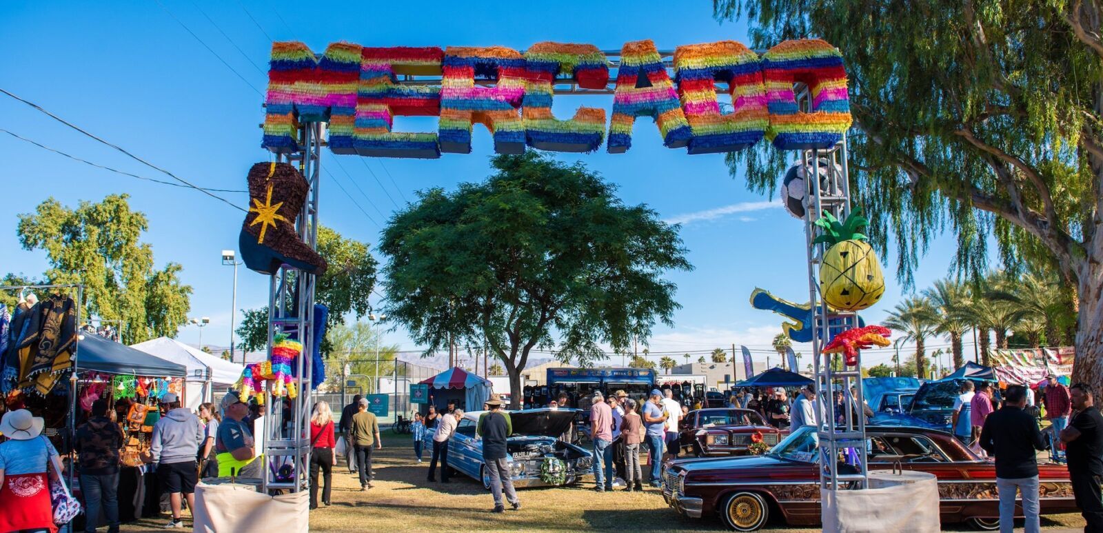 Colorful pinata-style MERCADO sign arch over lowrider cars and festival crowd at the Indio Tamale Festival