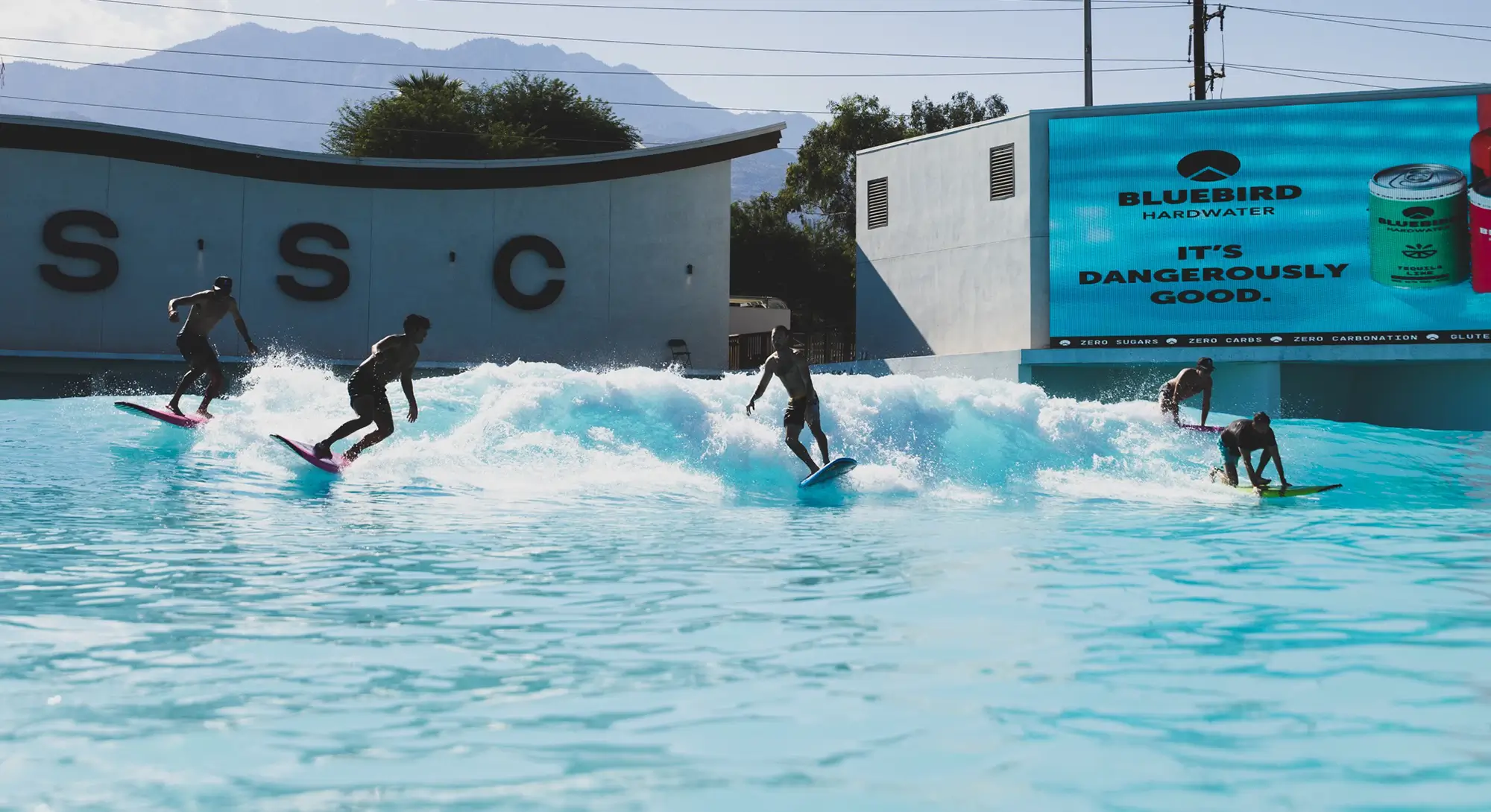 Multiple surfers riding the Wavegarden wave at Palm Springs Surf Club with the PSSC building and mountains behind them