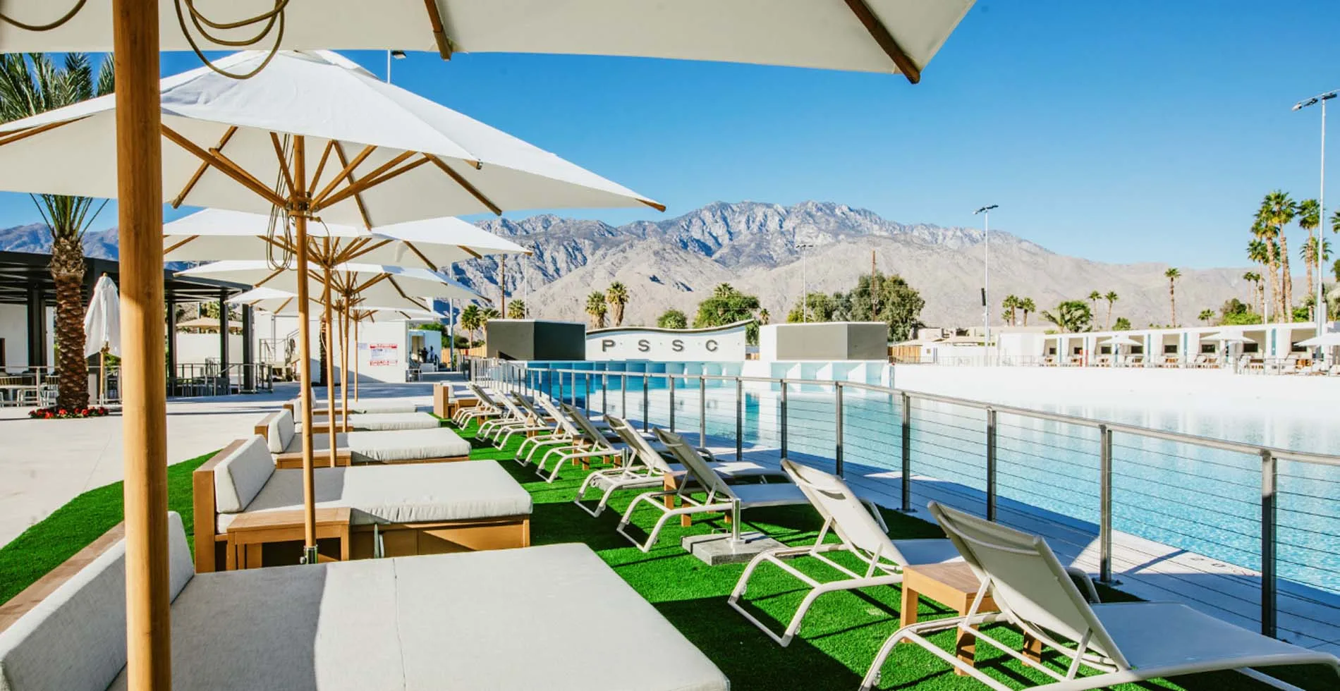 PSSC lounge area with white umbrellas and teak chairs facing the wave pool with San Jacinto Mountains behind