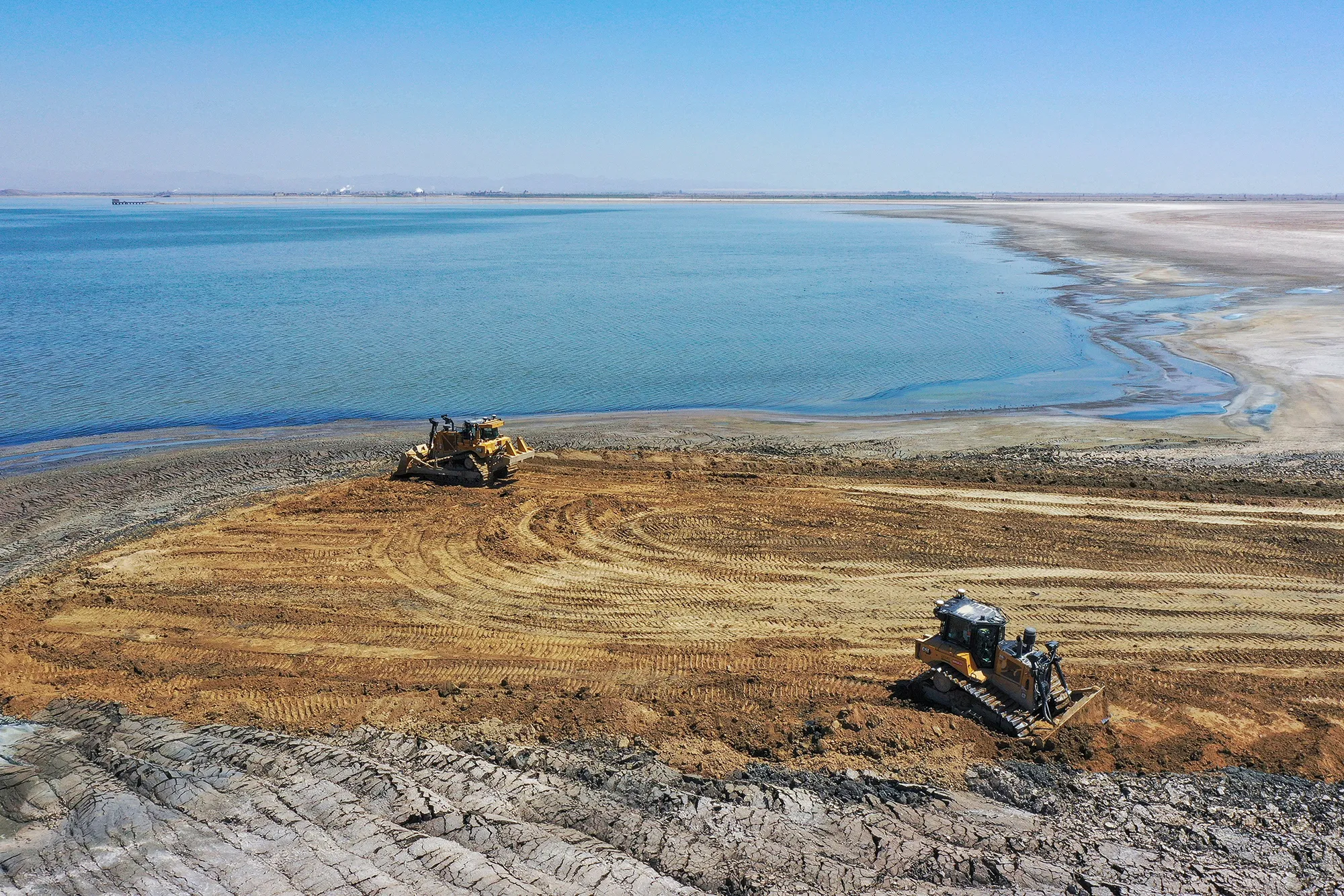 Aerial view of the Salton Sea shoreline with bulldozers on cracked earth and bright blue water, Southern California