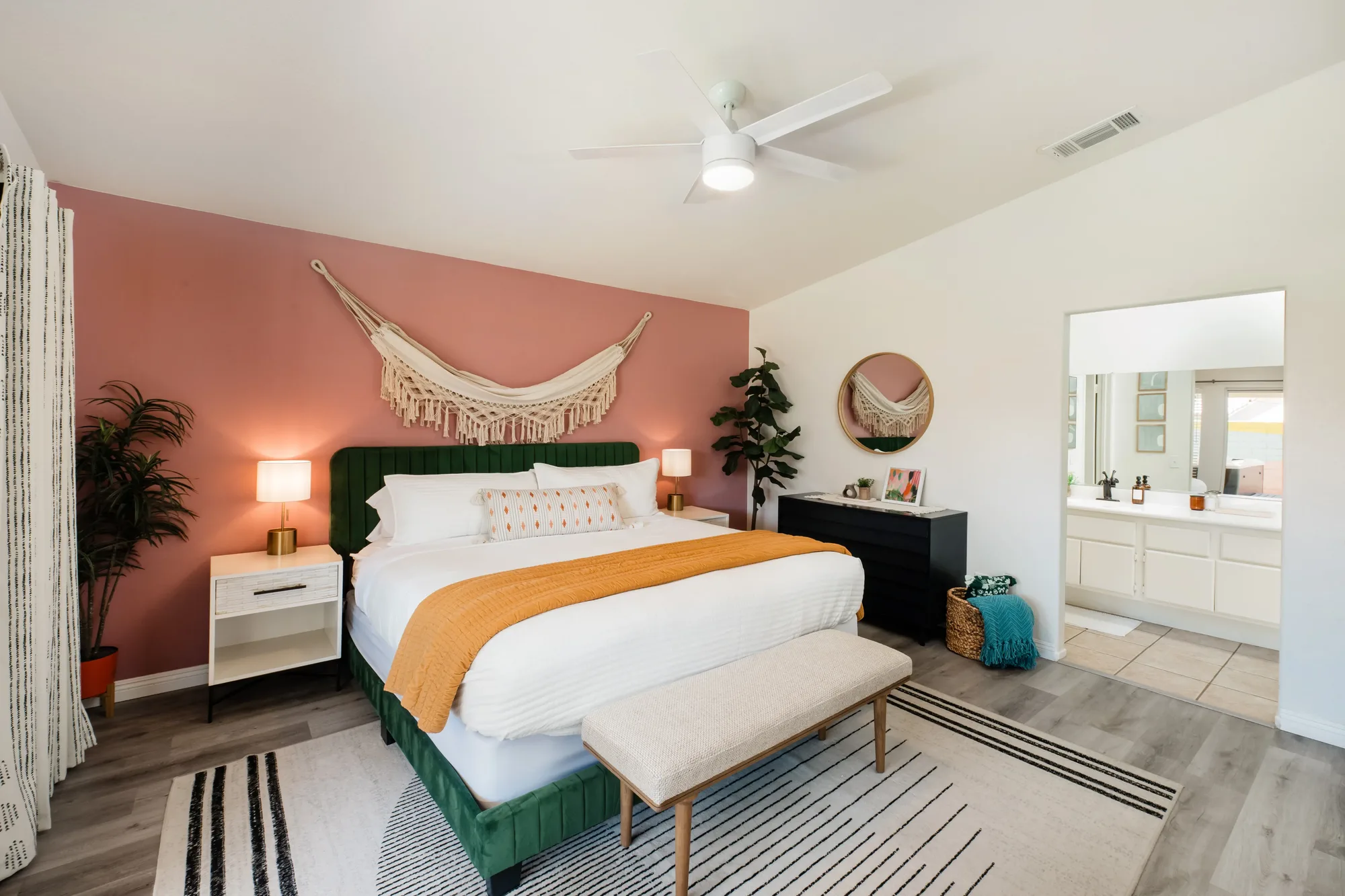 Cozy Cactus primary bedroom wide view with pink accent wall, green velvet king headboard, macrame art, and en-suite bathroom