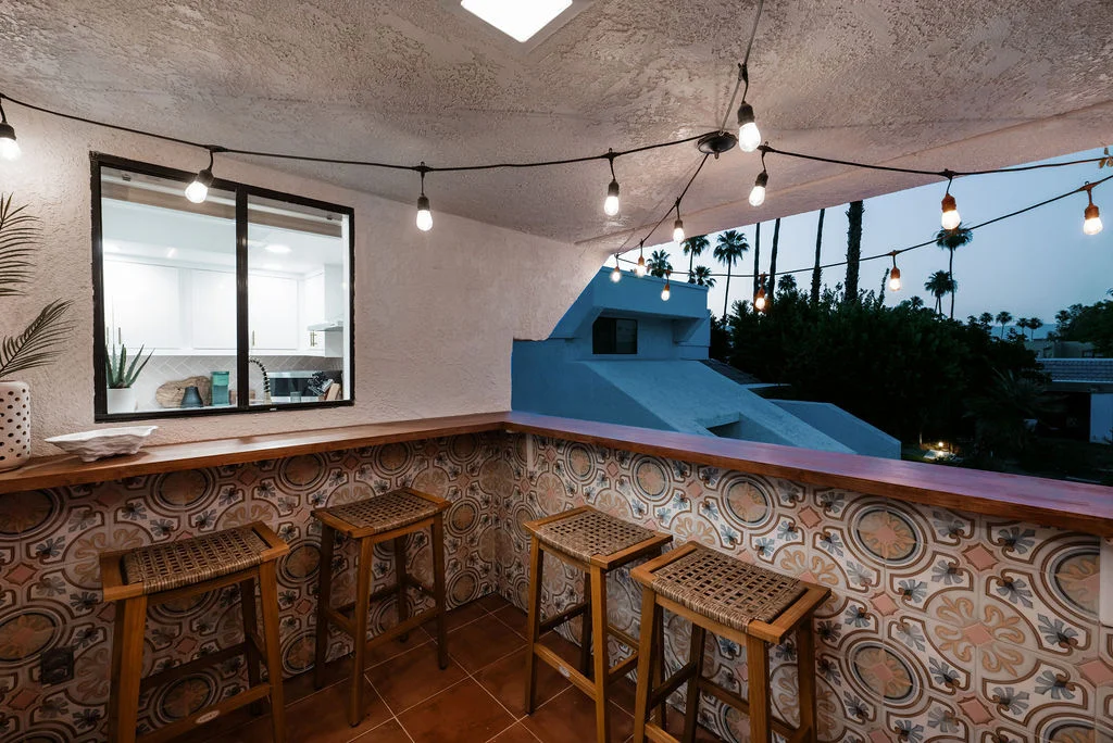 The Sundune Palm Springs balcony at dusk with string lights, cement tile, bar stools, and mountain view